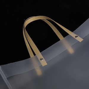K&ouml;nnte beinhalten: Eine transparente Plastikt&uuml;te mit zwei braunen Stoffgriffen. Die Griffe sind mit Metallnieten an der Tasche befestigt.