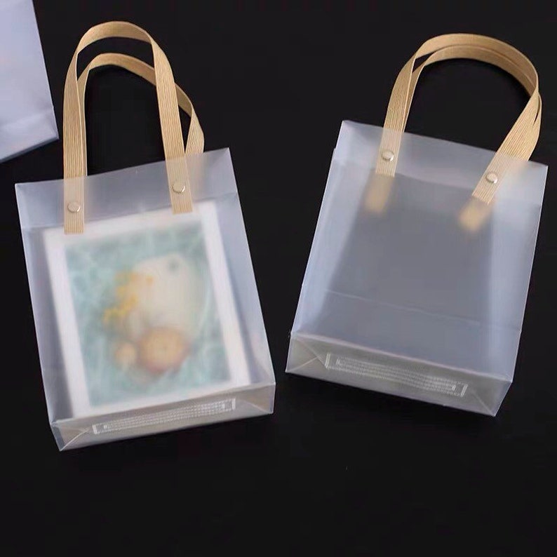 K&ouml;nnte beinhalten: Zwei transparente Plastikt&uuml;ten mit braunen Henkeln. Die Taschen haben eine matte Oberfl&auml;che und sind leer. Die Taschen befinden sich auf einer schwarzen Oberfl&auml;che.