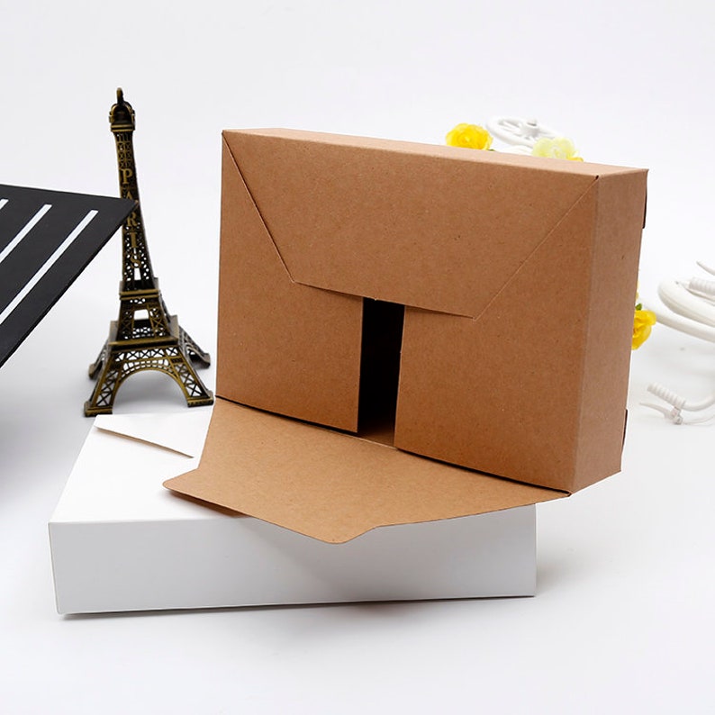 May include: A brown cardboard box with a unique flap design, partially open to reveal the interior. The box sits atop a white rectangular box. A miniature Eiffel Tower and a black and white object are in the background.