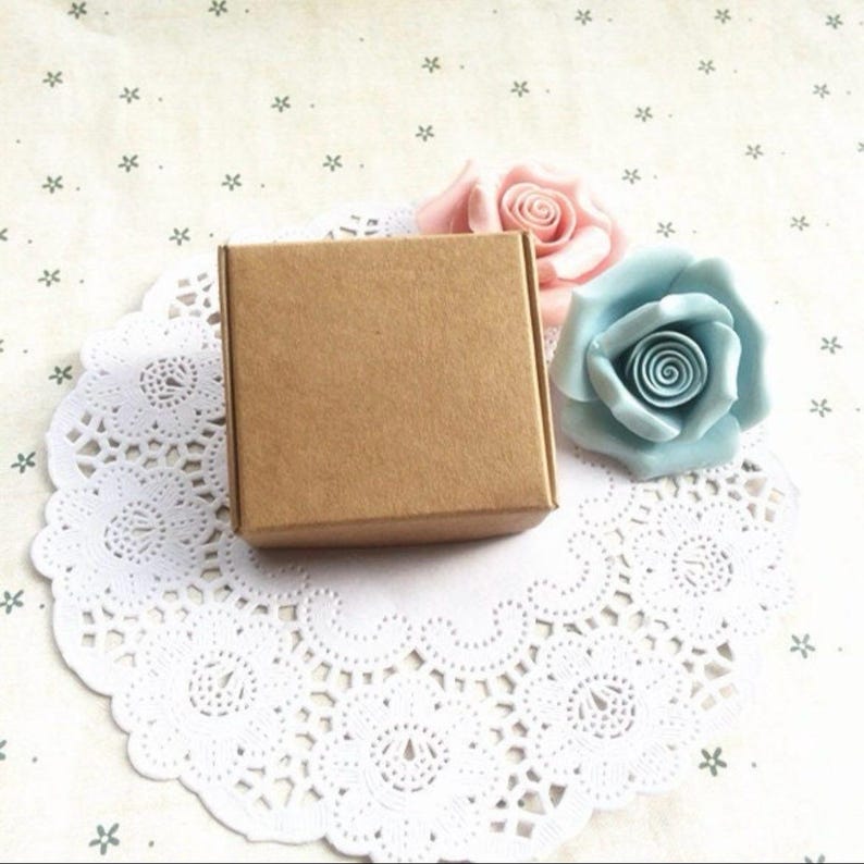 May include: A small, brown cardboard box with a square shape. The box is sitting on a white doily with two artificial flowers, one pink and one light blue.