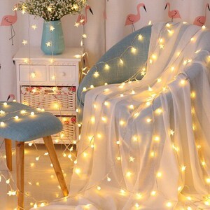 May include: A string of warm white fairy lights with star-shaped bulbs draped over a white sheer curtain and a blue chair. The lights are also on the floor and around a small wooden stool.