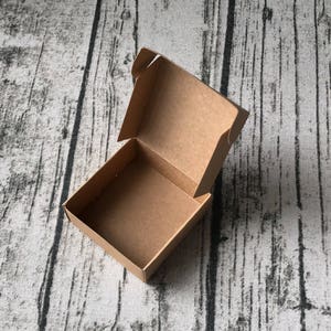 May include: An empty, open, brown cardboard box with a square base and a hinged lid. The box is sitting on a textured, grey and black surface.