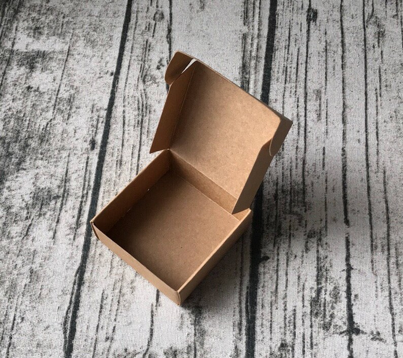 May include: An empty, open, brown cardboard box with a square base and a hinged lid. The box is sitting on a textured, grey and black surface.