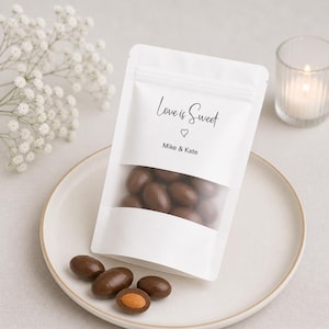 Op de afbeelding: Een witte zak met een doorzichtig venster gevuld met chocolade-amandelen. De zak heeft de tekst "Love is Sweet" en de namen "Mike & Kate". De zak ligt op een wit bord met verspreide amandelen en een brandende kaars.