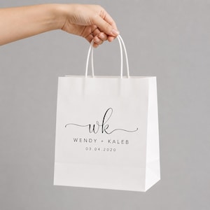 May include: A white paper gift bag with two white handles. The front features black printed text with the lowercase letters wk in a cursive script, followed by the names WENDY + KALEB and the date 03.04.2020 in a simple sans-serif font.