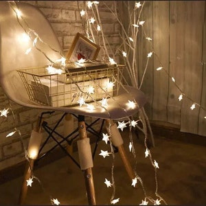 May include: White string lights with star-shaped bulbs draped over a white chair and a wooden branch. The lights are turned on and create a warm glow.