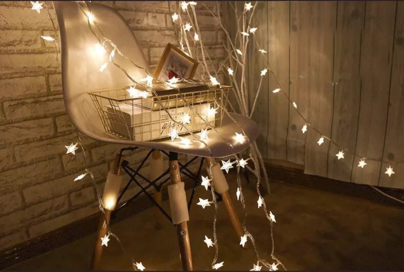 May include: White string lights with star-shaped bulbs draped over a white chair and a wooden branch. The lights are turned on and create a warm glow.