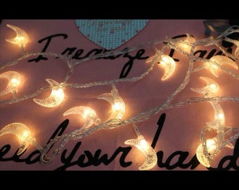 May include: A string of white fairy lights with crescent moon shaped bulbs. The lights are strung on a clear wire and are lit with a warm white glow.