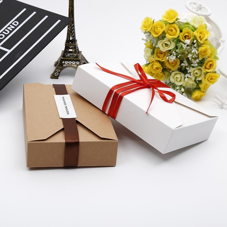 May include: Two gift boxes, one brown and one white, are displayed. The brown box is wrapped with a dark brown ribbon and a "HAND MADE" label. The white box is tied with a red ribbon. A bouquet of yellow roses and a miniature Eiffel Tower are in the background.