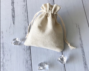 Könnte beinhalten: Eine kleine, cremefarbene Kordeltasche aus Naturstoff, mit drei klaren Quarzkristallen, die auf einer weißen Holzoberfläche verstreut sind.