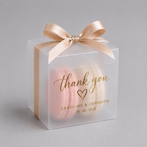 Puede incluir: Una caja de regalo cuadrada de plástico transparente atada con un lazo de satén color champán. El frente presenta texto en lámina dorada que dice thank you, un icono de corazón, CAROLINE & JOHNSON y 06.06.2032, con dos macarons visibles.