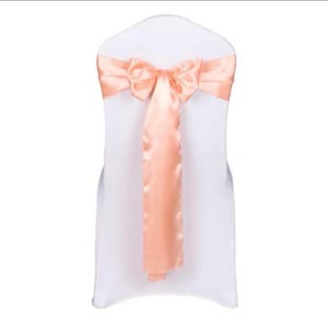 Puede incluir: Una faja de silla de satén melocotón atada en un lazo en una silla blanca. La faja mide aproximadamente 25 cm de ancho y 76 cm de largo.