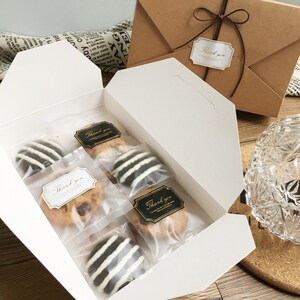 May include: A white gift box filled with individually wrapped cookies. Some cookies have white and dark chocolate stripes. The box also contains cookies with chocolate chips. "Thank you" stickers are on the cookie packaging and the brown envelope.