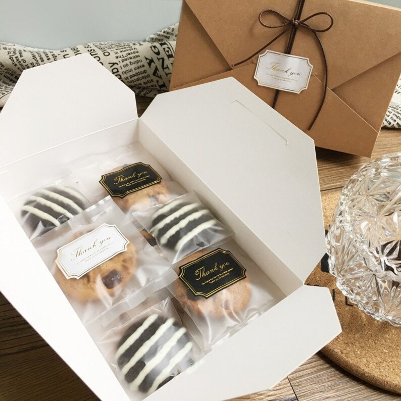 May include: A white gift box filled with individually wrapped cookies. Some cookies have white and dark chocolate stripes. The box also contains cookies with chocolate chips. "Thank you" stickers are on the cookie packaging and the brown envelope.
