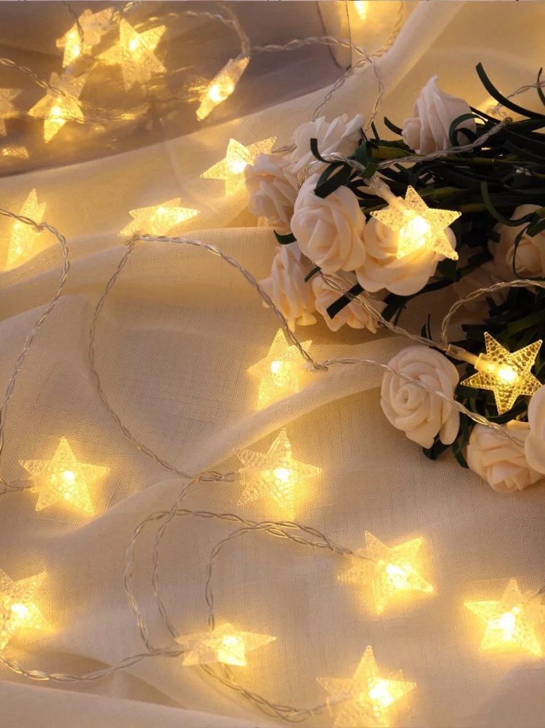 May include: A string of white fairy lights with star-shaped bulbs, illuminating a white fabric background. The lights are strung around a bouquet of white roses.
