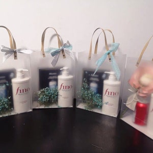 K&ouml;nnte beinhalten: Vier transparente Plastikt&uuml;ten mit Henkeln, jede mit einer Flasche Haarbehandlungsprodukt mit der Aufschrift "fino" und einem kleinen Strau&szlig; wei&szlig;er Blumen. Die Taschen sind mit einem blauen Band gebunden.