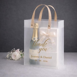 Op de afbeelding: Een transparante cadeauzak met een champagnefles en een witte doos met een strik. De tas heeft bruine handvatten en gouden tekst met de tekst "Thank you", namen en een datum. Een klein bloemstuk staat op de achtergrond.