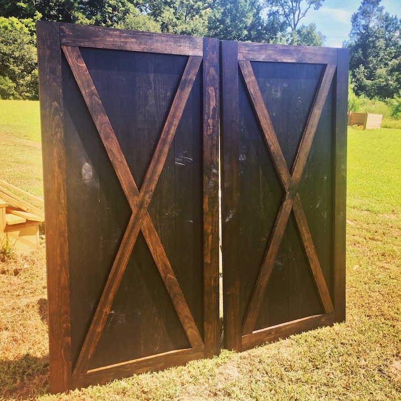 May include: A pair of tall, rectangular wooden doors featuring a dark brown stained finish and a prominent black X-shaped brace design. Each door is constructed with vertical planks and a thick outer frame, showcasing a visible wood grain texture.