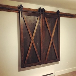 May include: A pair of dark brown wooden sliding barn doors featuring a prominent X-shaped brace design on each panel. The doors are mounted on a horizontal metal track system with black rollers, attached to a wooden header board above the door frame.