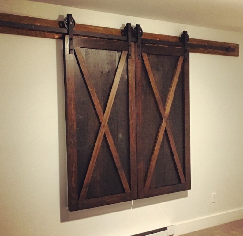 May include: A pair of dark brown wooden sliding barn doors featuring a prominent X-shaped brace design on each panel. The doors are mounted on a horizontal metal track system with black rollers, attached to a wooden header board above the door frame.