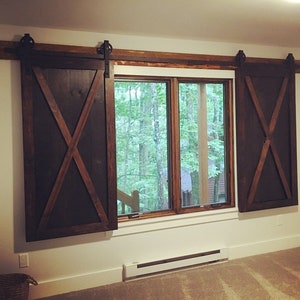 May include: Two dark brown wooden sliding shutters with a prominent X-shaped brace design mounted on a horizontal rail above a window. The shutters feature black metal hardware, including rollers and tracks, positioned against a white wall.