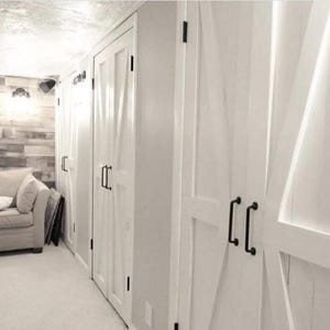 May include: A series of white double doors featuring a diagonal cross-brace design and matte black metal handles. The doors are installed in a row along a wall, providing access to storage spaces with a clean, bright finish and hardware.