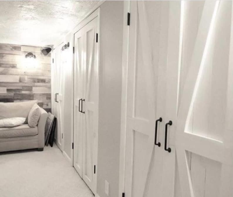 May include: A series of white double doors featuring a diagonal cross-brace design and matte black metal handles. The doors are installed in a row along a wall, providing access to storage spaces with a clean, bright finish and hardware.