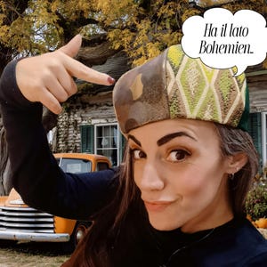 Può includere: Una donna indossa un cappello unico con un design patchwork di tessuti marroni, verdi e dorati. Il cappello ha una corona arrotondata e un bordo. L'immagine include il testo "Ha il lato Bohemien" in un fumetto.