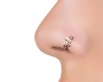 Può includere: Un anello per il naso in oro con un delicato design floreale e una piccola pietra preziosa trasparente. L'anello è indossato nella narice sinistra di una persona.