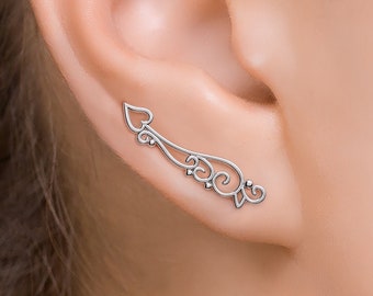 May include: Silver ear cuff with a delicate, ornate design featuring a heart shape and swirls. The cuff is shown on a person's ear.