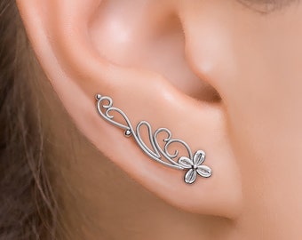 May include: Silver ear cuff with a delicate floral design. The cuff is curved and has a small flower at the end. It is a simple and elegant piece of jewelry.