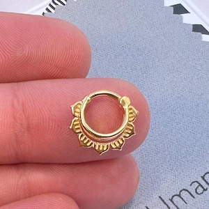 May include: Gold nose ring with a decorative design. The ring is shown on a finger and is packaged in a box with the brand name "Umanative" printed on it.