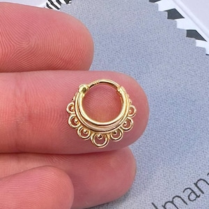 May include: Gold hoop nose ring with a decorative design around the hoop. The ring is on a finger.