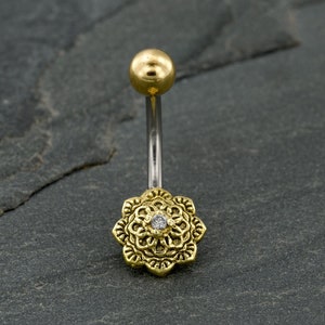 Könnte beinhalten: Goldener Bauchnabelring mit einem klaren Stein in der Mitte eines detaillierten Mandala-Designs.