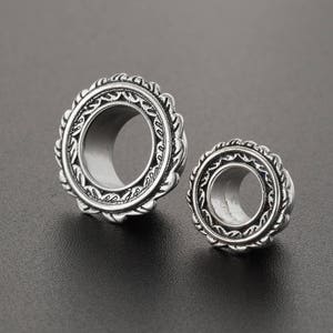 May include: Two silver ear gauges with intricate, ornate designs. The larger gauge has a diameter of approximately 1 inch, while the smaller gauge is about 0.75 inches in diameter.