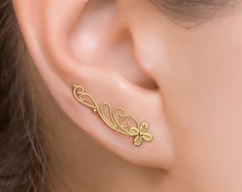 May include: Gold ear cuff with a delicate floral design. The cuff is curved and has a small flower at the end. It is a simple and elegant piece of jewelry.
