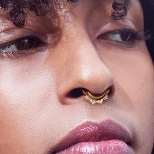 May include: Close-up of a person's face with dark curly hair and a gold nose ring. The nose ring has a decorative, sunburst-like design. The person has full lips and is looking off to the side.