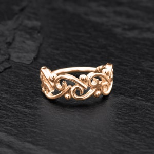May include: A gold-toned ring with a delicate, intricate design featuring swirling, floral-like patterns. The ring is made of metal and has a smooth, polished finish.