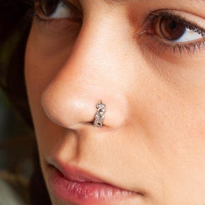 May include: Close-up of a person's face with a small silver nose ring. The person has dark brown hair and is looking off to the side.