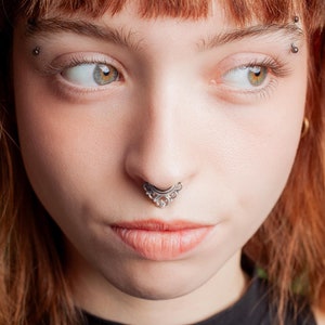 May include: A close-up portrait of a person with auburn hair and multiple facial piercings. The person has a septum piercing and eyebrow piercings. The background is blurred green foliage.