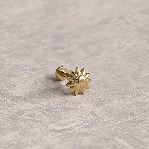 Może przedstawiać: Mały, złoty kolczyk w kształcie słońca. Kolczyk ma sztyft i dekoracyjny wzór promieni słonecznych. Kolczyk jest umieszczony na teksturowanej, jasnoszarej powierzchni.
