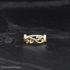 May include: A gold filigree ring with a delicate, intricate design. The ring is made of a thin, smooth metal and features a series of swirling, interconnected loops. The ring is displayed on a black, textured surface.