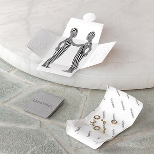 May include: A set of gold hoop earrings and a gold chain necklace with a small pendant, packaged in a white box with a black and white graphic design. The box is open and the jewelry is visible. The jewelry is on a white paper with the word "Umanative" printed on it.
