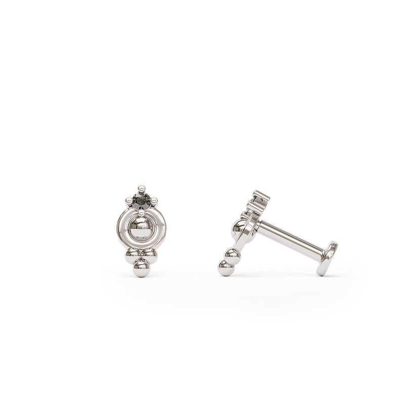 May include: A silver body jewelry piece with a black gem and a circular design. The jewelry is made up of multiple small balls and a larger ball in the center.