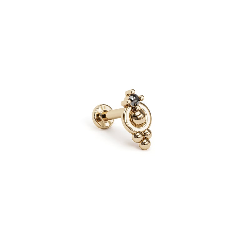 May include: Gold body jewelry with a small black gemstone and a circle design with three small balls hanging from the bottom.