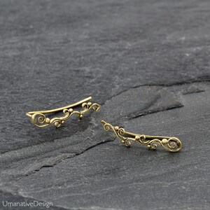 May include: A pair of gold ear cuffs with a delicate, floral design. The cuffs are made of brass and have a slightly curved shape.