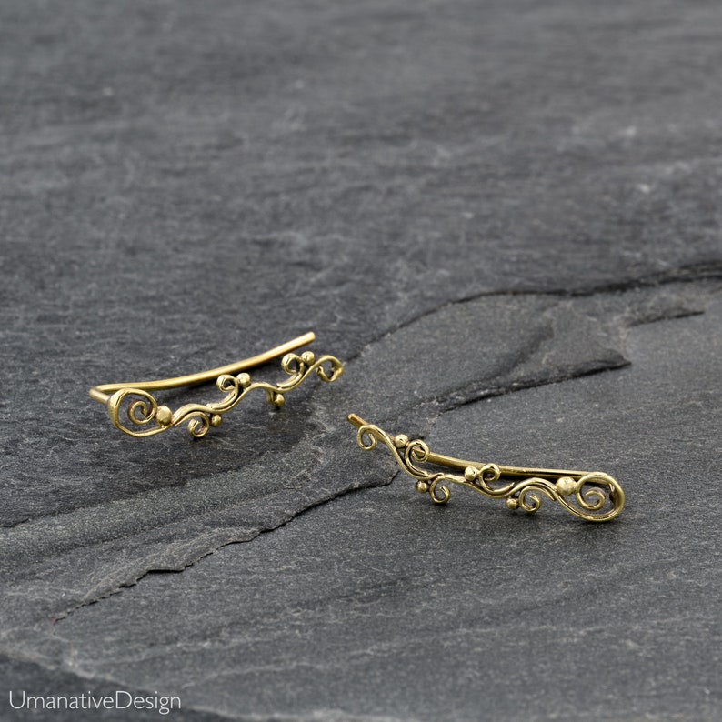 May include: A pair of gold ear cuffs with a delicate, floral design. The cuffs are made of brass and have a slightly curved shape.
