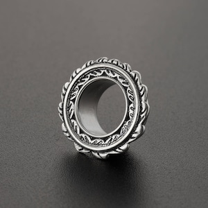 May include: Silver-toned metal ear gauge with an intricate, ornate design. The gauge has a circular shape with a large hole in the center.