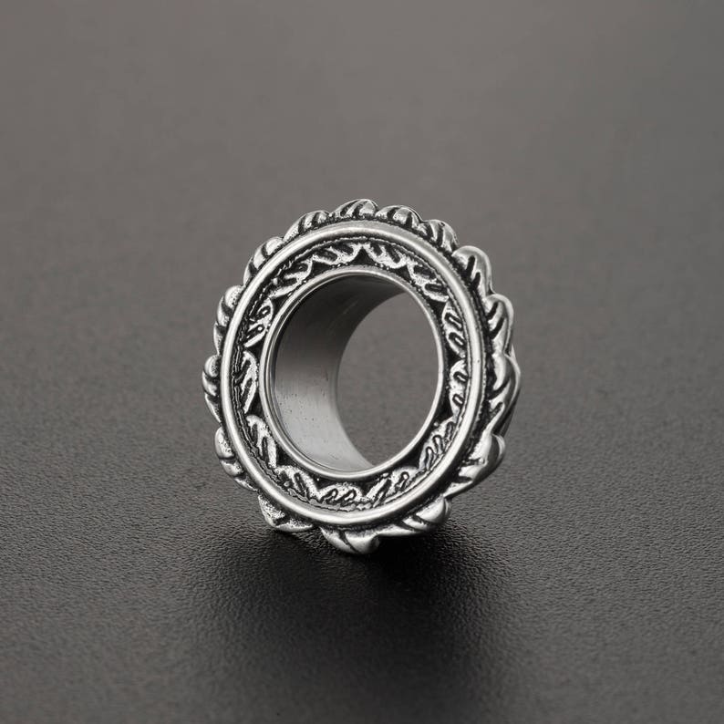 May include: Silver-toned metal ear gauge with an intricate, ornate design. The gauge has a circular shape with a large hole in the center.