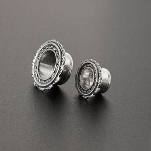 May include: Two silver ear gauges with an ornate design. The larger gauge has a diameter of approximately 1 inch, and the smaller gauge has a diameter of approximately 1/2 inch.